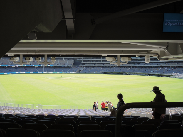 Eine Gruppe von Menschen vor einem großen Stadion, mit einigen Personen am Boden, Stühlen auf der rechten Seite und einem Bildschirm mit Text in der oberen rechten Ecke.