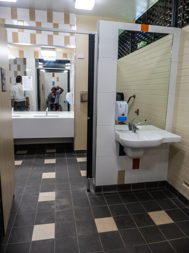 Ein Badezimmer mit Waschbecken, Spiegel und Deckenleuchten, mit einer Person im Spiegel reflektiert, die eine Kamera hält.