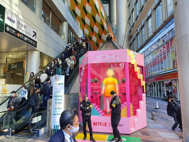 Eine Gruppe von Menschen steht vor einem Gebäude mit Glaswänden und Texttafeln, einige tragen Masken, mit einer Rolltreppe im Hintergrund; ein pinker Kasten in der Mitte zeigt Schaufensterpuppen und trägt den Text "Netflix's new Netflix booth in Tokyo."