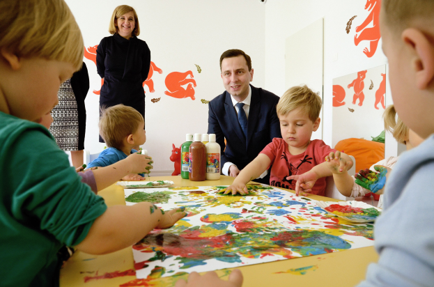 Kinder malen an einem Tisch mit Farbe an den Händen, Papieren und Flaschen, beaufsichtigt von einer lächelnden Frau, mit Gemälden an der Wand im Hintergrund.