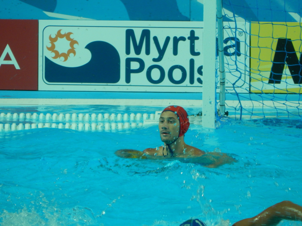 Ein Wasserballspieler mit Mütze in einem Pool mit einem Netz auf der rechten Seite und einer Werbetafel im Hintergrund.