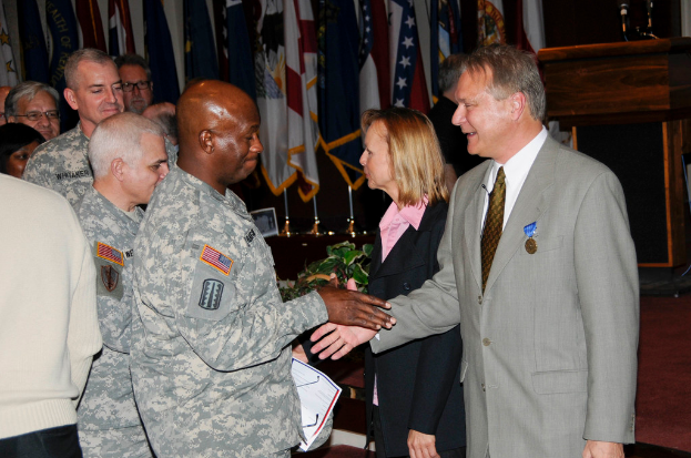 Eine Gruppe von Menschen, die mit zwei Personen Hand in Hand stehen, Flaggen im Hintergrund, ein Podium mit Mikrofon und eine Topfpflanze in der Nähe.