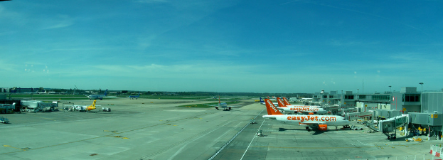 Eine Flughafen-Szene mit Flugzeugen, Geb├Ąuden, Masten, Fahrzeugen und B├Ąumen, mit einem Himmel ├╝ber und einer Stra├če darunter, und einem Zaun im Vordergrund.