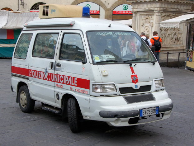 Ein weißer Van fährt auf einer Straße mit einer Gruppe von Menschen in der Nähe, die Taschen tragen und auf der rechten Seite eine Hütte zu sehen ist, mit Gebäuden und Metallstangen im Hintergrund.