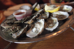 Ein Teller mit Meeresfrüchten in einer Reihe, eine Tasse Sauce in der Mitte, zwei Zitronenstücke daneben und im Hintergrund sichtbare Teller auf einem Tisch.