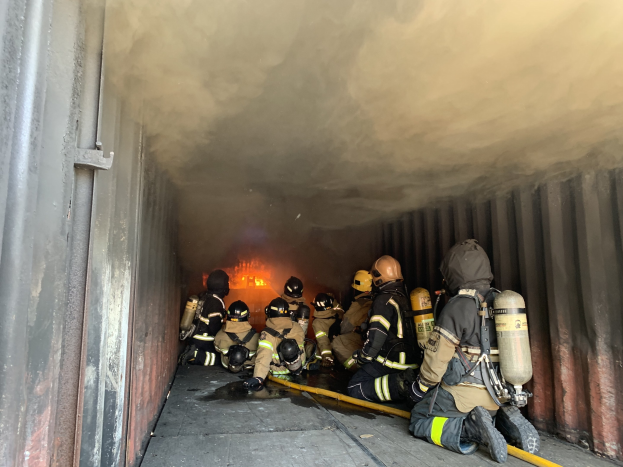 Feuerwehrleute in Schutzausrüstung mit Sauerstoffflaschen arbeiten in einem raucherfüllten Tunnel, einer hält einen Schlauch, mit Flammen im Hintergrund und einer Wand auf der linken Seite.
