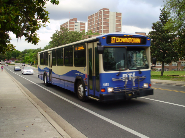 Ein blauer und weißer Shuttlebus fährt eine Straße mit hohen Gebäuden entlang, mit ein paar Menschen auf dem Bürgersteig links und Bäumen, Pfählen und einem klaren blauen Himmel im Hintergrund.