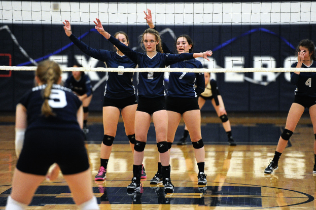 Drei Mädels bei einem Volleyballspiel auf dem Feld stehend, mit erhobenen Händen, ein Netz davor und ein Zaun im Hintergrund.