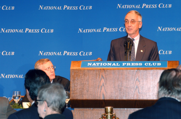 Ein Mann in einem Blazer, Krawatte und Brille spricht in ein Mikrofon an einem Pult mit einer Tafel vor einem sitzenden Publikum, mit einer Werbung im Hintergrund.
