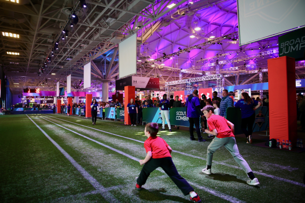 Zwei junge Jungs spielen Fußball auf einem Feld, umgeben von einer Gruppe von Menschen, mit Flaschen und anderen Gegenständen auf der rechten Seite, einem Zaun, Bannern, einem Anzeigebildschirm, Metallrahmen, einem Dach mit Deckenleuchten und Metallstangen im Hintergrund.