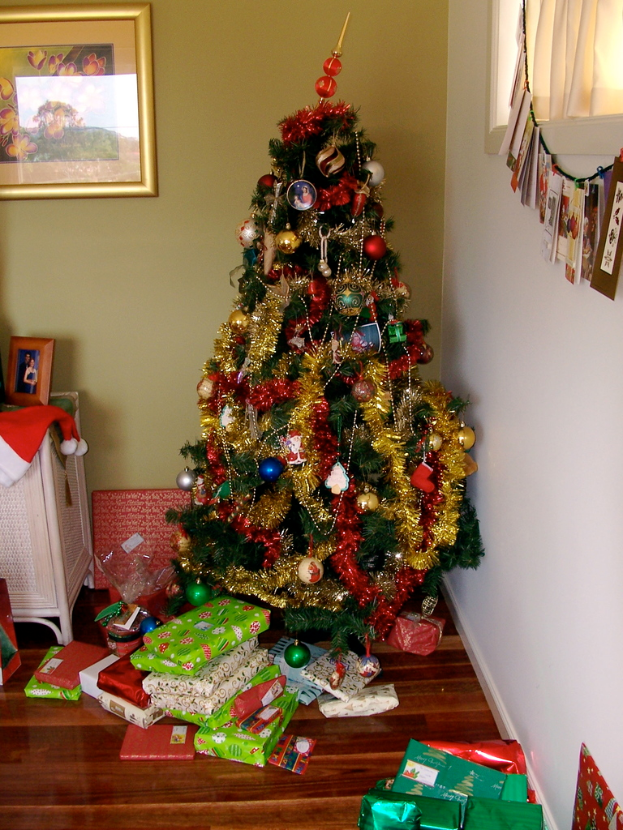 Ein geschmÃ¼ckter Weihnachtsbaum rechts mit Geschenken darunter, ein Foto Rahmen an der linken Wand und ein Vorhang am rechten Fenster.