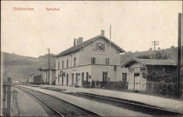Schwarzes und weißes Foto einer Bahnstation in Dielkirchen, Deutschland, mit einem Zug auf den Gleisen, umgeben von Häusern, Strommasten, Bäumen, Menschen und Hügeln im Hintergrund unter einem klaren Himmel, mit dem Text "Dielkirchen, Bahnhof, Deutschland" oben und unten.