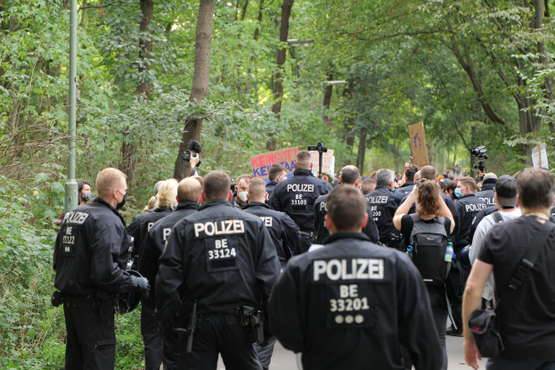 Eine Gruppe von Polizisten in schwarzen Uniformen geht eine von Bäumen gesäumte Straße entlang, einige tragen Taschen und Kameras, mit einem Pfahl im Hintergrund.