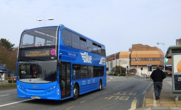Ein blauer Bus mit Text darauf fährt auf einer Straße, mit einem Mann, der rechts auf dem Gehweg geht und einem Schild in der Nähe. Links parkt ein schwarzes Auto hinter einem Zaun mit Gras, Bäumen und einer Wand. Im Hintergrund gibt es ein Gebäude, Bäume und ein weiteres Schild, unter einem bewölkten Himmel.