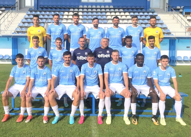 Mannergruppe auf einem Fußballfeld, einige sitzen auf Bänken und andere stehen auf dem Rasen, mit Tribünen und architektonischen Elementen im Hintergrund.