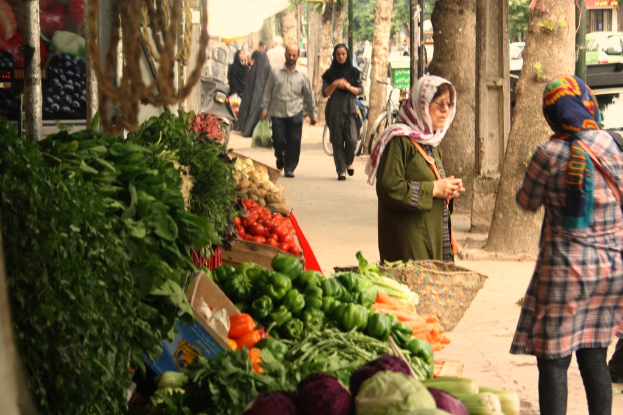 Ein belebter Gemüsemarkt mit verschiedenen Produkten wie Paprika, Tomaten und Kartoffeln auf der linken Seite, Menschen, die auf der rechten Seite stehen und gehen, sowie Bäume und Gebäude im Hintergrund.