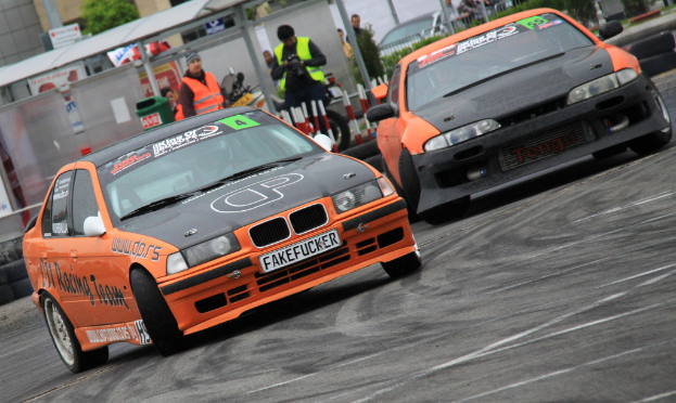 Zwei Autos drifteten auf einer Straße, mit einigen Mönner hinter einem Zaun mit einem Fahrrad und einer Bushaltestelle mit Geböuden im Hintergrund.