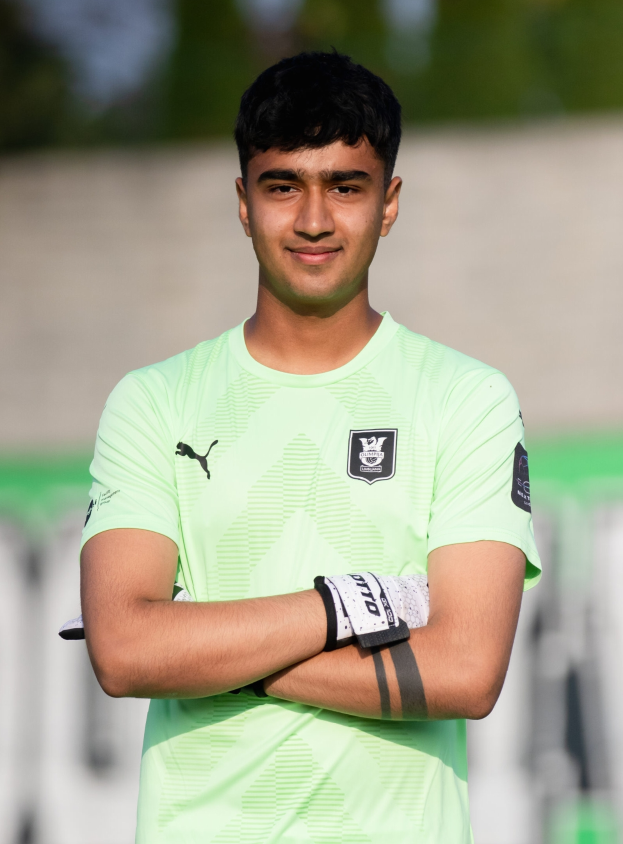 Ein junger Mann in einem grünen T-Shirt steht auf einem Fußballfeld und schaut selbstbewusst in die Kamera, während der Hintergrund unscharf ist und den Fokus auf ihn lenkt.