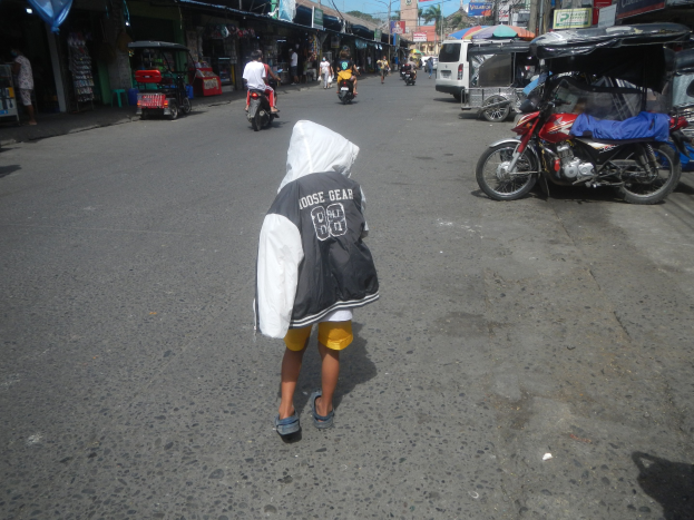 Ein junger Junge geht eine Straße voller Motorräder und anderer Fahrzeuge entlang, flankiert von Gebäuden mit Schildern, unter einem klaren blauen Himmel mit Bäumen und Pfählen im Hintergrund.