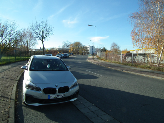 Ein BMW 2er Active Tourer 218d xDrive M Sport fährt auf einer Straße, umgeben von Bäumen, Pfählen, Laternen, Gebäuden und einem Zaun, mit einem klaren blauen Himmel im Hintergrund.