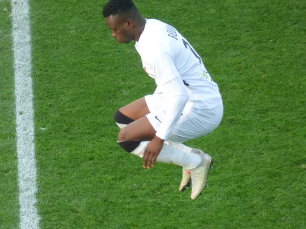 Ein Mann in weißer Uniform und Fußballstiefeln kniet auf einem Fußballfeld, wobei auf der linken Seite der Bildes eine weiße Linie zu sehen ist.