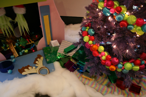 Drinnen steht ein geschmückter Weihnachtsbaum mit Lichterkette und Schmuck auf der rechten Seite, umgeben von Geschenkboxen und verschiedenen dekorativen Gegenständen, mit einer Hütte und Tüchern im Hintergrund.