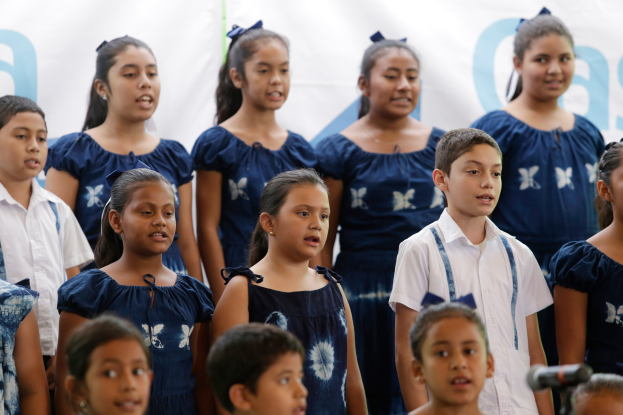 Eine Gruppe von Kindern steht vor einem Mikrofon mit einem Schulchor-Banner im Hintergrund, die scheinbar singen.
