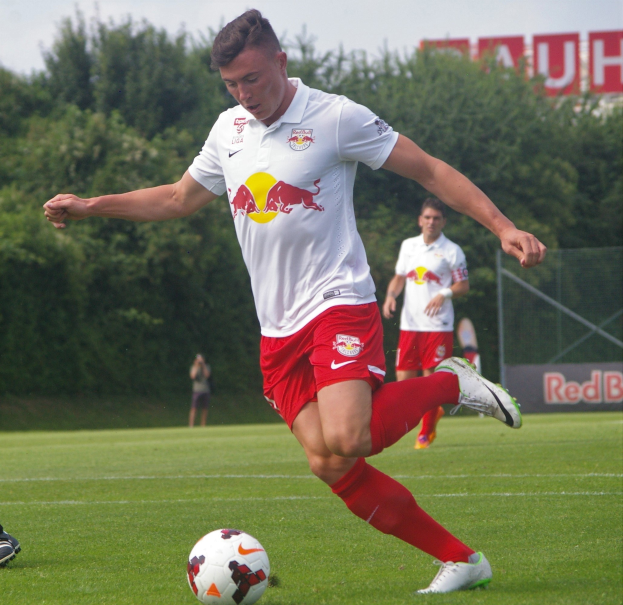 Ein professioneller Fußballspieler schießt einen Ball auf einem Rasenfeld mit Bäumen und einem klaren blauen Himmel im Hintergrund, wobei ein "RB Leipzig"-Schild sichtbar ist.