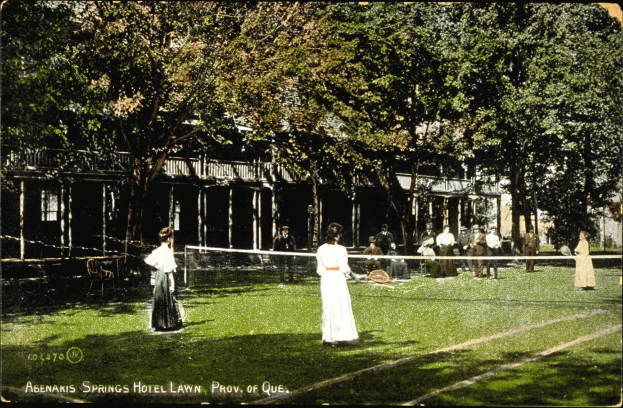 Schwarz-weiß-Foto einer Gruppe von Menschen, die Tennis auf einem Rasenplatz vor dem Abenakis Springs Hotel Lawn in Provo, Quebec spielen, wobei einige auf Stühlen sitzen, ein Netz, Bäume und ein Gebäude im Hintergrund.