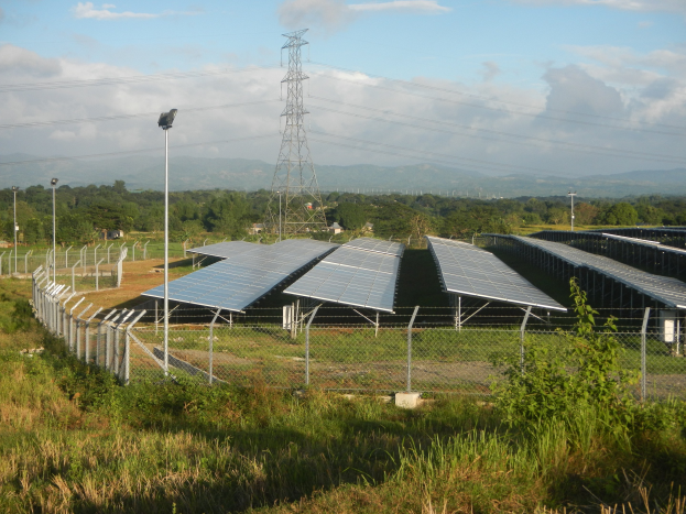Ein Solarpanelfeld, das von einem Zaun umgeben ist, mit Gras, Pflanzen und Bäumen und einem Übertragungsturm mit Drähten im bewölkten Himmel.