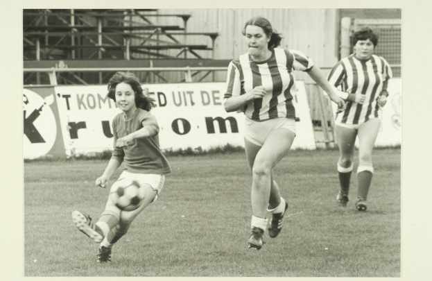 Drei Frauen in Schwarz-Weiß-Foto beim Fußballspielen auf einem Feld, tragen Fußballtrikots und Turnschuhe, mit Bannern, Stangen und einer Wand im Hintergrund.