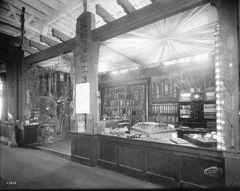 Schwarz-weiß-Foto eines alten Apothekenladens mit einem zentralen Tresen, auf dem verschiedene Gegenstände ausgebreitet sind, Regalen im Hintergrund gefüllt mit Flaschen und Artikeln und Text unten, der darauf hinweist, dass es sich um eine Apotheke handelt.