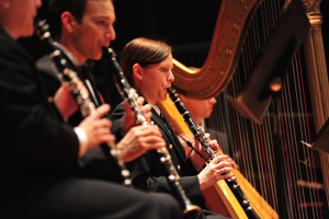 Eine Gruppe von Menschen sitzt auf einer Bühne und spielt Klarinetten, mit einem Ständer im Vordergrund und einem dunklen Hintergrund, der eine lebendige Atmosphäre schafft.