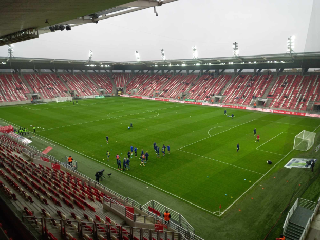 Ein Fußballfeld in einem Stadion mit Menschen, Stühlen, Geländern, Treppen und Lampen, unter einem sichtbaren Himmel.