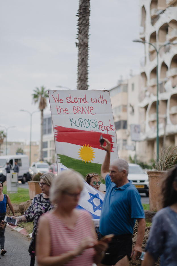 Eine Gruppe von Menschen marschiert nachts eine Straße entlang, mit einem Schild, auf dem "Wir stehen zu den tapferen kurdischen Menschen" steht, mit einem auf der Straße geparkten Van, Straßenlaternen, Bäumen, Gebäuden und einem klaren blauen Himmel im Hintergrund.