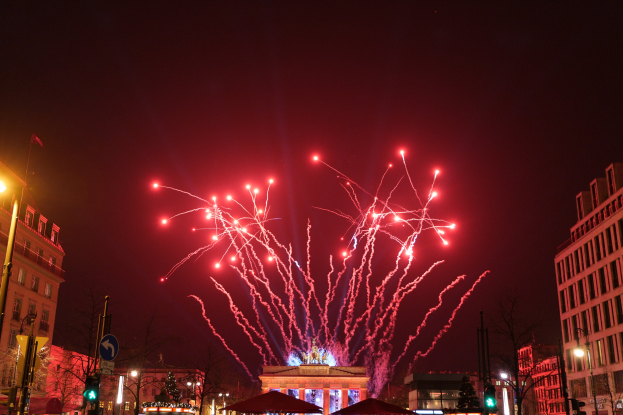 Eine belebte Stadtstraße an einem Neujahrsabend in Berlin, mit Gebäuden, Bäumen, Laternenmasten, Verkehrszeichen, Zelten, Menschen und einem prächtigen Feuerwerk am Himmel.