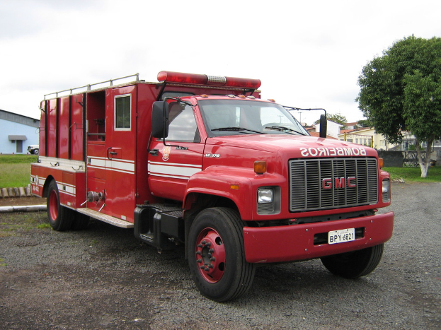 Kraftfahrzeuge auf einer Straße mit Bäumen, Gras, Gebäuden und einem bewölkten Himmel im Hintergrund.