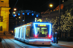 Ein Straßenbahnzug fährt nachts eine Straße entlang, mit Menschen, die auf dem Bürgersteig rechts von der Bahn stehen, und Gebäuden, Polen, Lichtern und dekorativen Lichtern im Hintergrund, die eine festliche Atmosphäre schaffen.