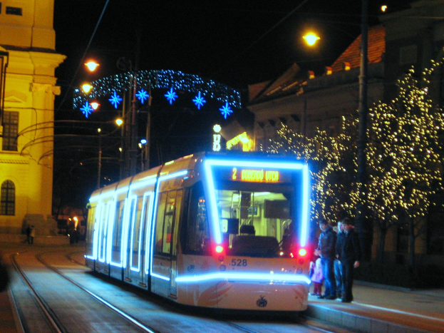 Ein Straßenbahnzug fährt nachts eine Straße entlang, mit Menschen, die auf dem Bürgersteig rechts von der Bahn stehen, und Gebäuden, Polen, Lichtern und dekorativen Lichtern im Hintergrund, die eine festliche Atmosphäre schaffen.