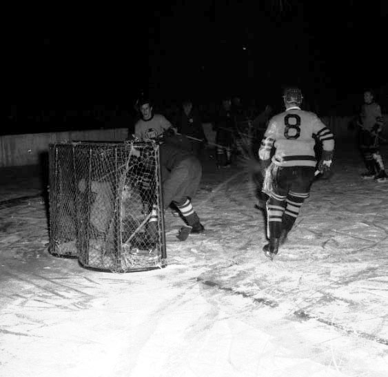 Eine Gruppe von Männern, die Hockey auf einem Eisplatz spielen, mit einem Netz im Vordergrund und einer Wand im Hintergrund, dargestellt in Schwarz-Weiß.