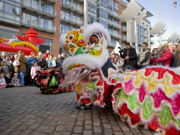 Ein lebendiges chinesisches Neujahrsfest in Amsterdam mit einem Löwen tanzen im Vordergrund mit einer Menge von Menschen, einige halten Kameras, die sich um es versammelt haben, vor Buildings, Laternenmasten und einem klaren blauen Himmel.