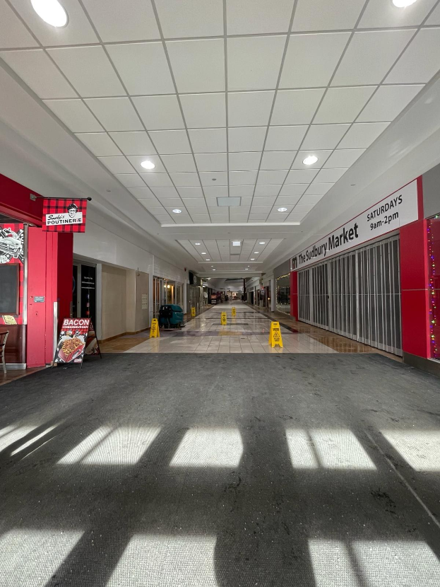 Innenseite eines Einkaufszentrums mit Deckenbeleuchtung, einem Stuhl und einer Tafel, die links " Sydbury Market - Vorher und Nachher " zeigt, und verstreuten Gegenständen rechts und auf dem Boden.