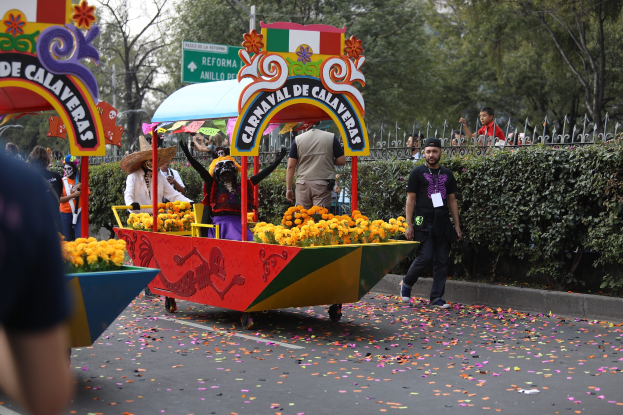 Eine Gruppe von Menschen auf einem blumen geschmückten Festwagen bei einem Umzug, die verschiedene farbige Kleider und Hüte tragen, mit Schildern, einem Metallzaun, Bäumen und einem bewölkten Himmel im Hintergrund.