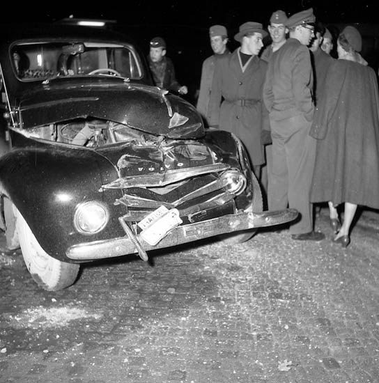 Schwarz-weißes Bild eines schwer beschädigten Autos am Straßenrand mit einer Gruppe von Menschen, die die Szene begutachten.