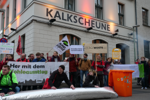 Eine Gruppe von Menschen hält Schilder und Plakate vor einem Gebäude, während im Vordergrund zwei Personen sitzen und rechts ein Müllcontainer steht, bei einer Demonstration in Deutschland, mit Gebäuden, Fenstern, Lichtern und Schildern im Hintergrund.