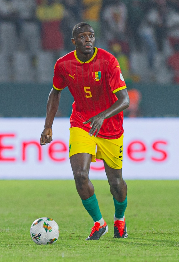 Ein Mann in rot-gelber Uniform spielt Fußball auf einem Feld, trägt Turnschuhe und hat eine Tafel im Hintergrund sowie einige Zuschauer auf den Rängen.