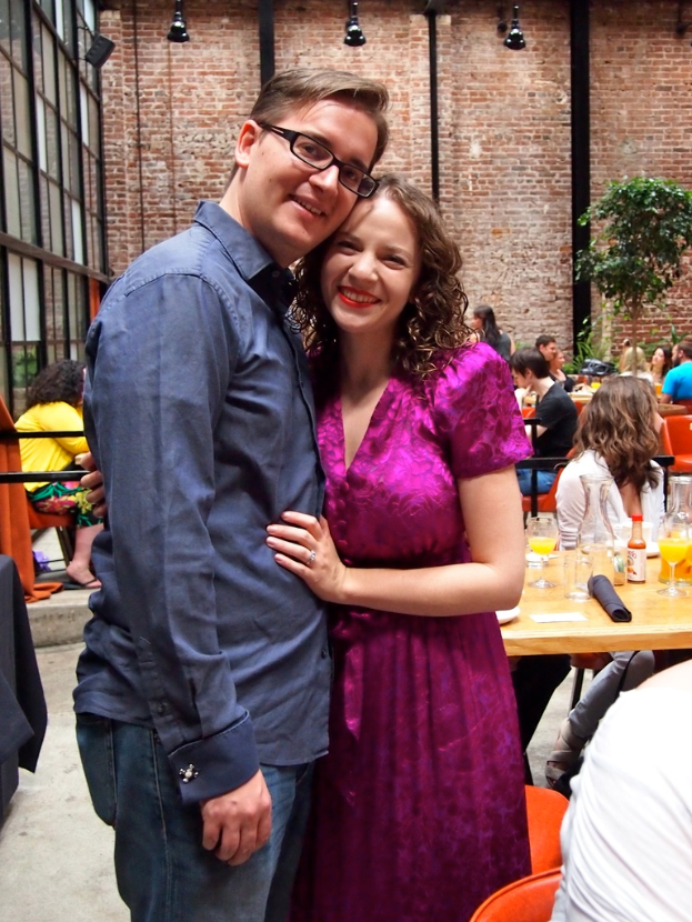 Ein Mann und eine Frau lächeln vor einer Backsteinwand mit Lichtern, dahinter sitzen Menschen an Tischen mit Gläsern, Flaschen, Säften und einer Pflanze.