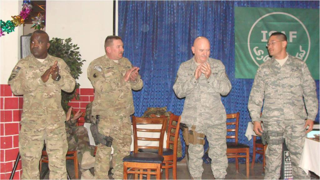Vier Personen stehen, drei klatschen, eine sitzt auf einem Stuhl neben einer Pflanze, mit einem Banner an einem Vorhang im Hintergrund, alle tragen Militäruniformen, Stühle und ein Tisch sind sichtbar.