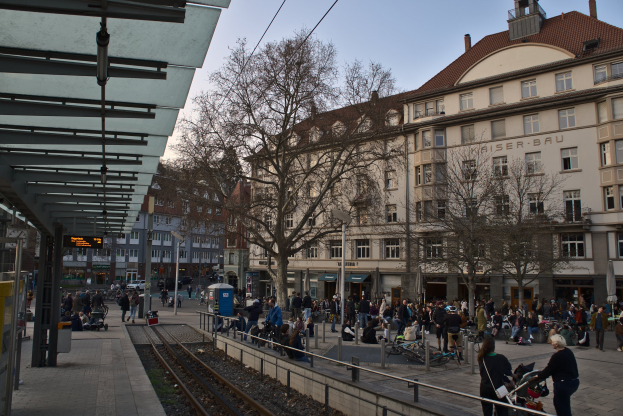 Eine belebte Stadtstraße mit einem Bahnhof auf der linken Seite, umgeben von Gebäuden, Bäumen und Laternen, mit Menschen, die gehen, auf Bänken sitzen oder in der Nähe des Bahnhofs stehen, unter einem sichtbaren Himmel.