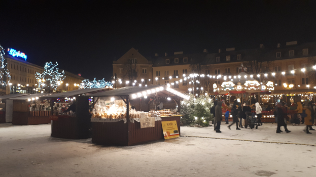 Lebhafter Weihnachtsmarkt mit Menschen, Buden mit Lichtern, Pflanzen, Bäumen, Gebäuden und Schildern auf einer verschneiten Nachtstraße unter einem bewölkten Himmel.